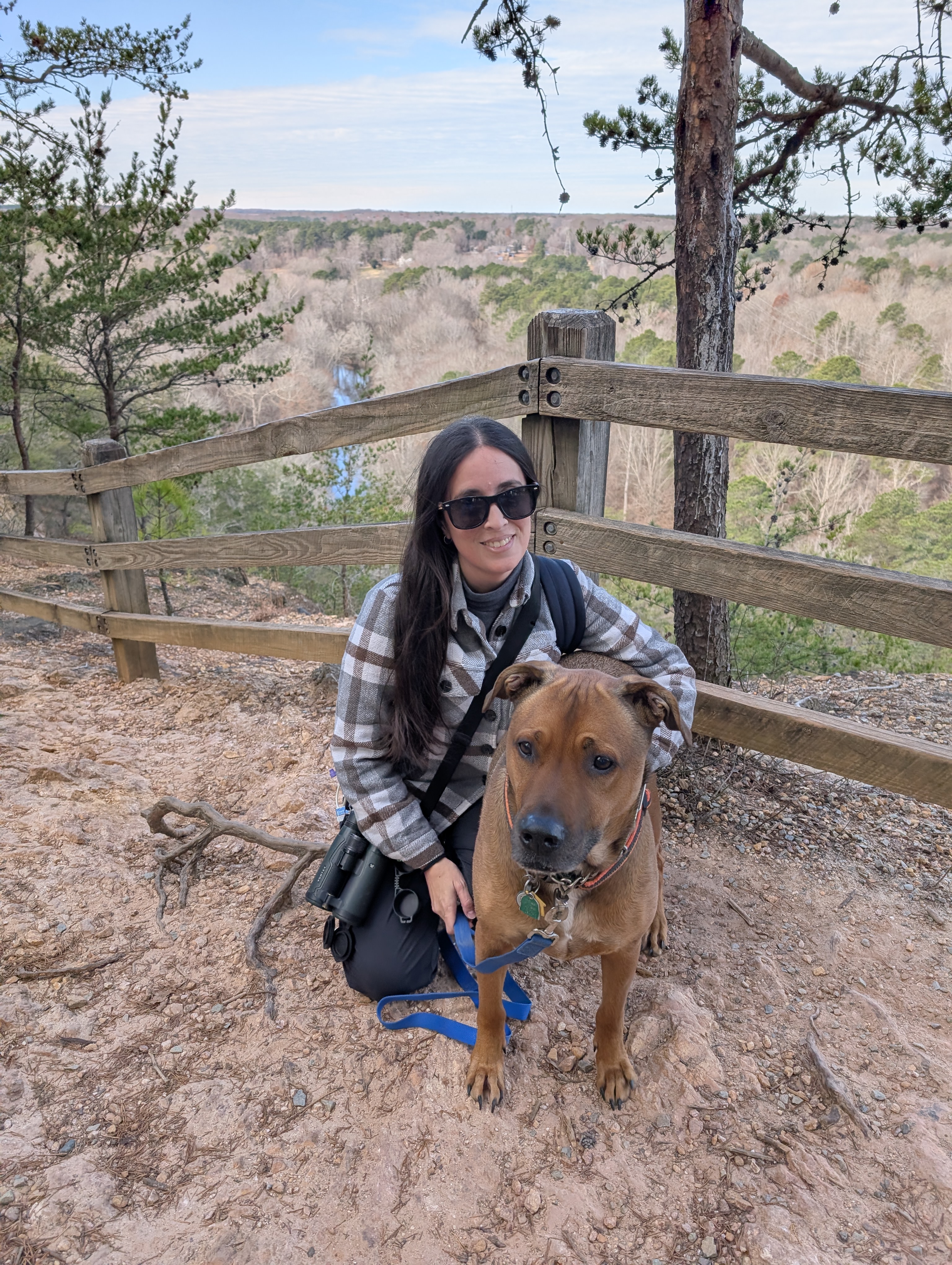 A woman in sunglasses kneels beside a brown dog on a trail overlooking a wooded valley. 
      She's smiling, and the scene feels peaceful and serene.