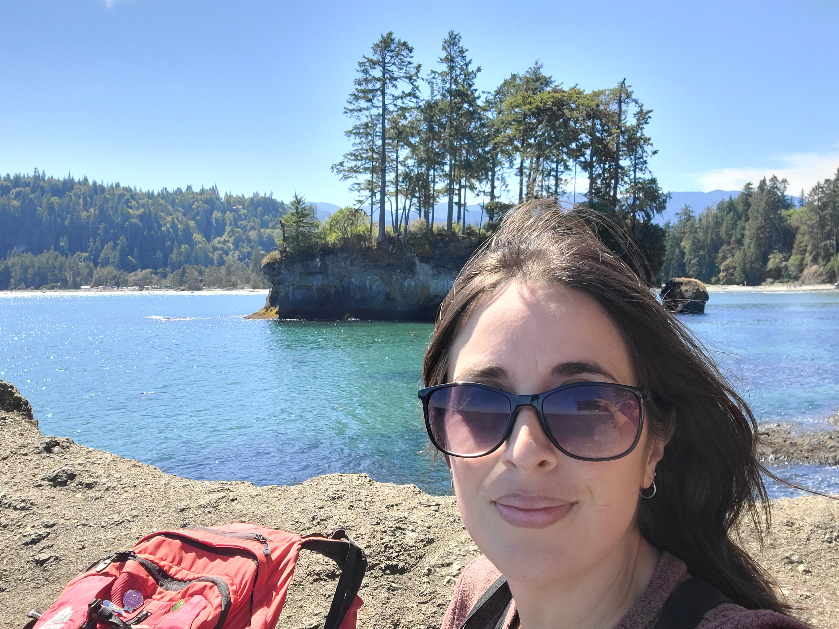 A woman with long dark hair and sunglasses smiles near a scenic coastline. Clear blue water, dense green trees 
      on an island, and a bright sky create a calm atmosphere. A red backpack is beside her.