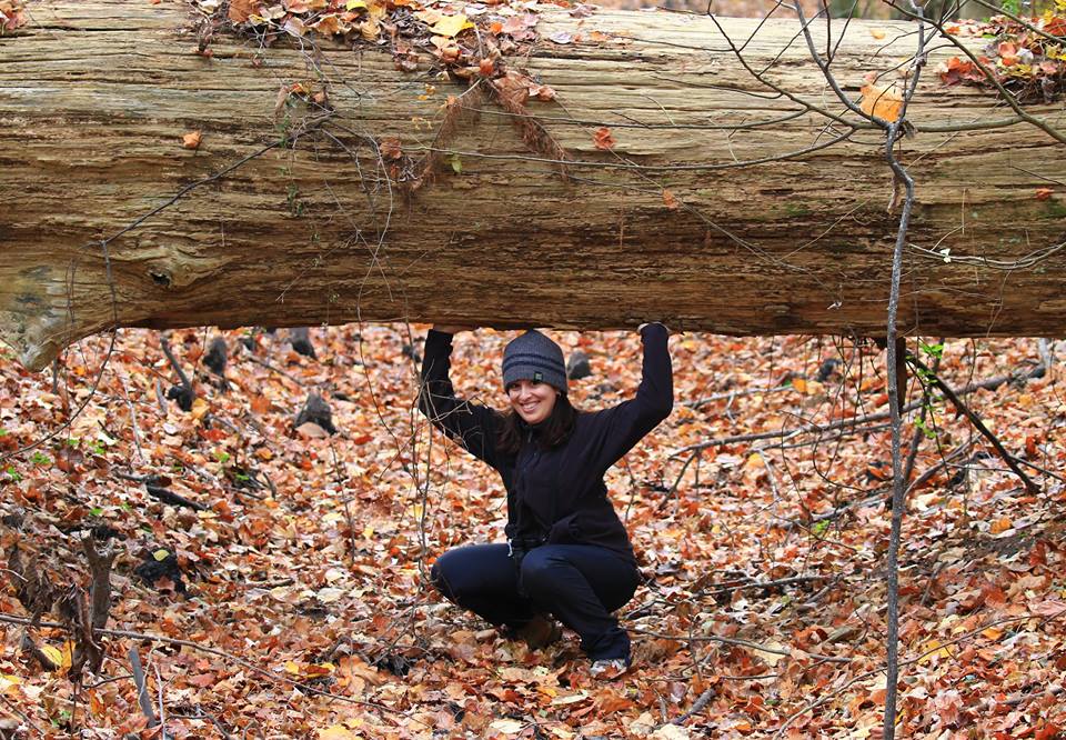 A person crouches beneath a fallen tree trunk in a forest, 
      surrounded by autumn leaves. They wear a black outfit and a gray beanie, smiling confidently.